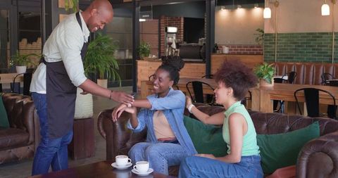 African american barista serving two friends on leather sofa in modern coffeehouse lounge