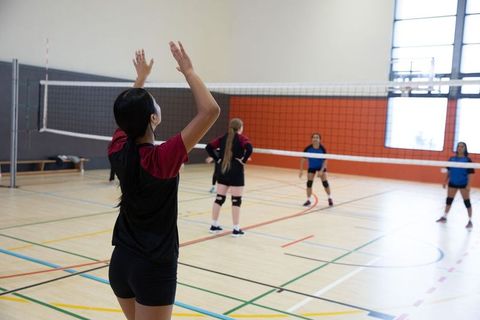 Teenage volleyball players serving and preparing defense on indoor gym court junior match