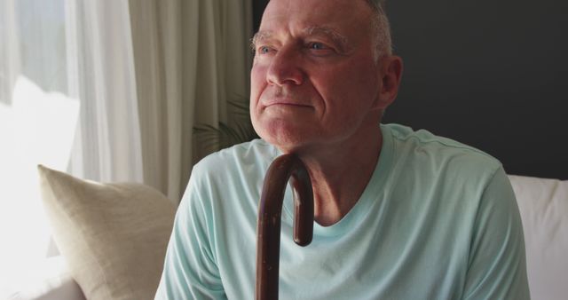A senior man is holding a cane and sitting thoughtfully at home. The image captures a moment of reflection and calm. It is ideal for use in contexts related to aging, retirement planning, healthcare, eldercare services, or lifestyle pieces focusing on senior citizens. This visual evokes a sense of serenity and can complement stories about personal reflection and the lives of elderly individuals.