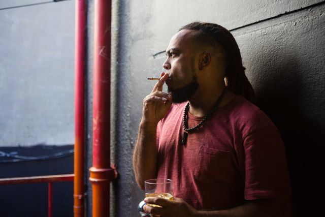 Man standing at bar entrance, smoking cigarette and holding glass of whisky. Suitable for themes of nightlife, relaxation, urban lifestyle, and casual evening activities.
