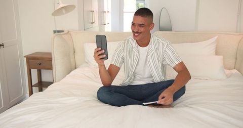 Young man sitting on bed using smartphone indoor relaxation