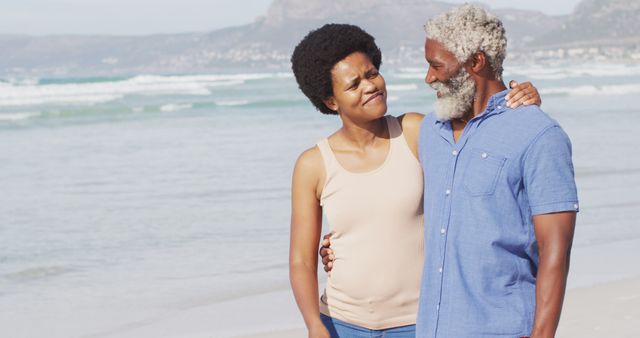 Senior African American couple walking arm in arm on beach, waves in background. They are smiling and enjoying the outdoors, creating a warm and loving atmosphere. Perfect for use in projects related to retirement, senior living, love, or holiday travel.