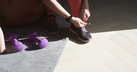 Adult female tying running shoes during home workout with purple dumbbells and smartwatch