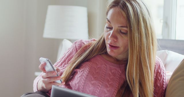 This image of a woman using a smartphone and tablet while sitting on a couch is ideal for projects related to technology, digital communication, and modern lifestyle. It highlights the integration of devices in everyday life and can be used in articles, blog posts, and advertisements focusing on technology usage, home environments, and multitasking in daily routines.