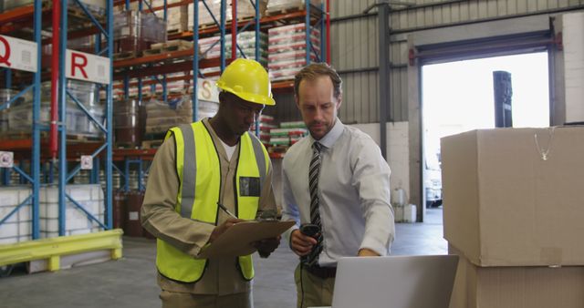 Two warehouse workers collaborating, one holding clipboard and the other operating digital equipment, showcasing teamwork in a logistic environment. The person on the left wears a hard hat and safety vest, emphasizing safety precautions. Suitable for themes related to logistics, warehouse operations, teamwork, inventory management, and workplace safety.