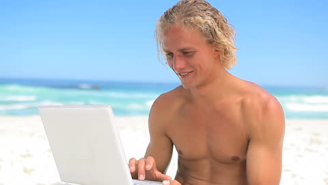 Smiling Blonde Man Using Laptop on Sunny Beach