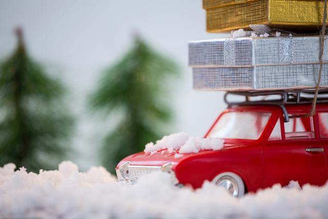Toy car carrying stacked Christmas presents on snowy ground with blurred Christmas trees in background. Ideal for holiday greeting cards, festive decorations, winter-themed advertisements, and Christmas promotions.