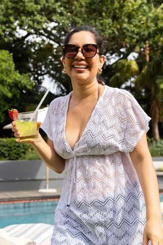 Smiling woman holding cocktail at poolside wearing lace cover-up and sunglasses enjoying vacation