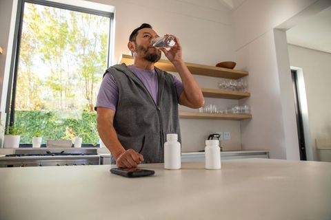 Mature asian man drinking water while taking supplements at modern kitchen island