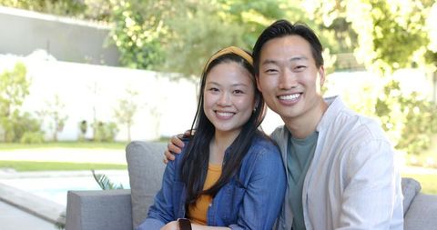 Asian couple smiling and sitting on backyard sofa by pool, woman wearing wrist device