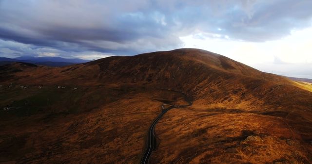 A winding road cuts through the expansive, undulating landscape of golden-brown hills under a dramatic sky. The scene captures the rugged beauty of a remote area, inviting viewers to imagine a journey through this serene yet untamed environment.