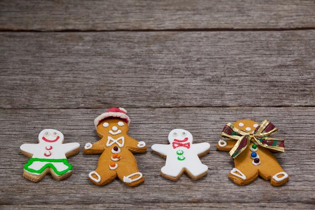 Four decorated gingerbread cookies with varied designs are arranged in a row on a rustic wooden table. The decorations include icing in various colors, buttons, and bows, making them an ideal subject for holiday-themed projects. This image can be used for holiday cards, blogs, festive advertising, recipes, or family-oriented activities.