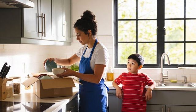 Woman unpacking dishes in modern kitchen with child watching by sunny window and sink