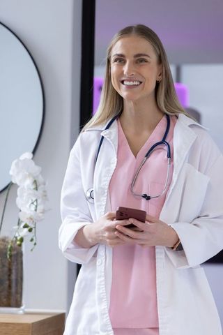 Smiling female clinician holding smartphone in modern clinic wearing pink scrubs and white coat