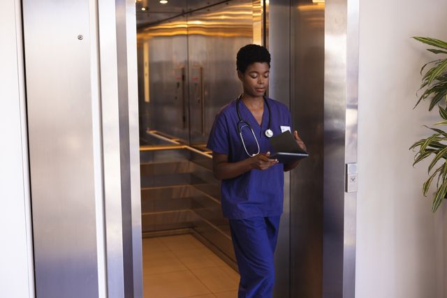 Smiling african american female doctor looking at tablet, walking out of elevator. Medical services, hospital and healthcare concept.