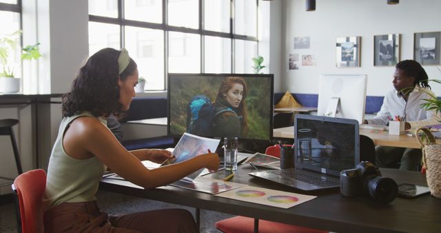 This image shows young creative professionals working in a modern office environment. One woman is examining photographs while another person is working at a computer in the background. The workspace is equipped with laptops, cameras, and design materials, emphasizing a collaborative and technological working atmosphere. This image is ideal for articles, websites, and marketing materials focusing on modern workspaces, creative industries, teamwork, and business environments.