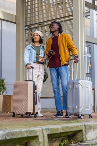 Diverse travelers waiting with suitcases at urban station holding coffee, smartphone