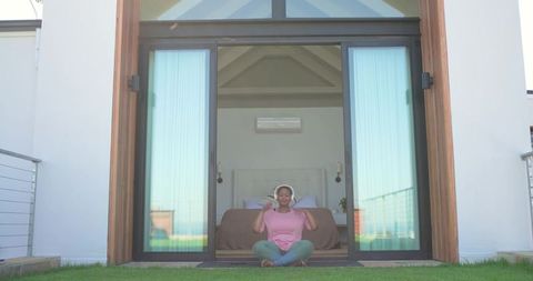 Woman sitting cross-legged on lawn listening with headphones at modern sliding doors