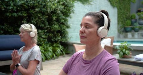 Two mature women meditating outdoors by pool wearing headphones for guided relaxation