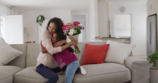 Mother and daughter sitting on a light gray sofa in a bright and cozy living room, sharing a warm hug. Daughter is holding a bouquet of flowers. Scene captures the joy and affection between mother and child, making it perfect for use in family, parenting blogs, Mother’s Day promotions, and advertisements celebrating family bonds.