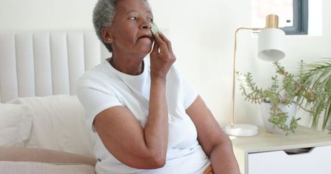Senior african-american woman applying facial pad while sitting on bed in pastel bedroom