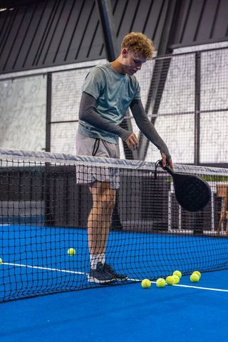 Young man scooping padel balls at net on indoor blue court during practice session