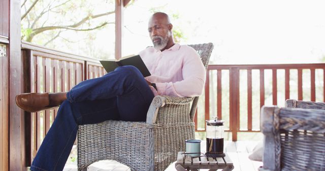 Mature man sitting on wicker chair on porch, reading book, enjoying peaceful leisure time. A cup of coffee and French press on side table. Useful for lifestyle, retirement, relaxation, and senior well-being concepts.