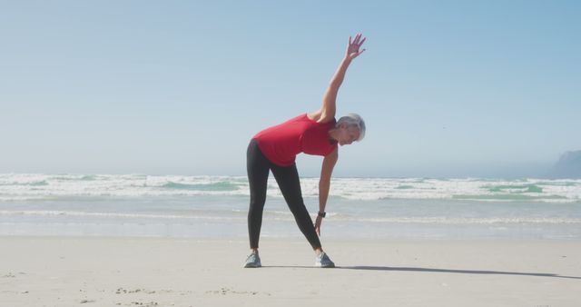 Senior woman wearing red top, performing a stretching routine on a sunny beach. Ocean waves in the background create a serene atmosphere. Useful for promoting fitness programs, healthy living, senior health tips, and active outdoor lifestyles.