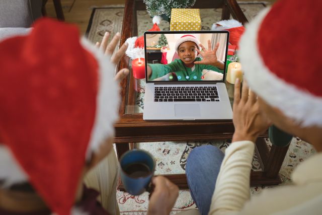 A diverse couple wearing Santa hats sit in a festive living room, having a joyful video call with an energetic and happy African American boy on a laptop screen. This image is perfect for illustrating themes of modern holiday celebrations, remote communication with loved ones, and embracing digital technology to stay connected during festive seasons. Ideal for blogs, articles, and marketing materials focusing on global holidays, virtual family gatherings, and Christmas traditions.