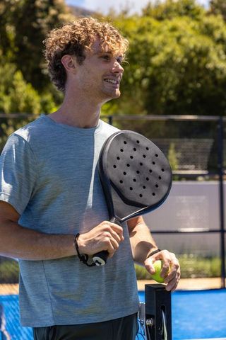 Smiling padel player holding perforated racket and ball on blue outdoor court during practice