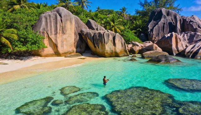 Wading male angler casting in turquoise tropical cove amid granite boulders and reef