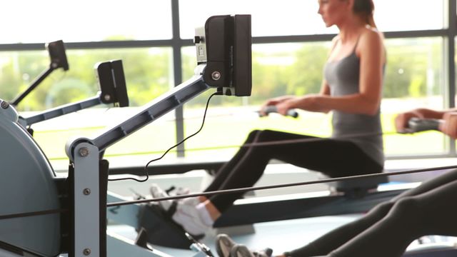 Women are exercising on rowing machines in a well-lit modern gym with large windows. The focus is on maintaining a healthy lifestyle and fitness routine. Ideal for content related to exercise routines, gym promotions, and active lifestyles.