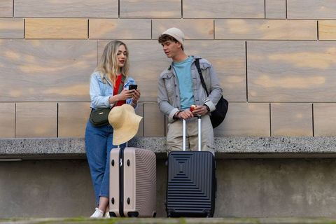 Young travelers standing with suitcases comparing smartphone near sandstone wall