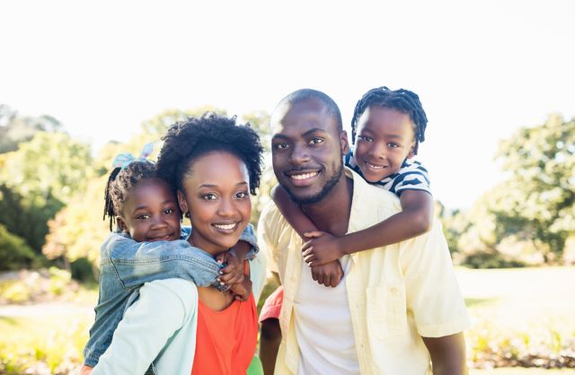Smiling African American family of four posing happily outdoors in a park. The parents are standing close to each other with their children on their backs. Ideal for use in advertisements and campaigns promoting family values, outdoor activities, parenting tips, and nature excursions.