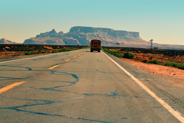 This image shows an old school bus traveling on a long, empty desert highway. The barren landscape includes patches of greenery and cracked pavement, with a grand mountain range prominently visible under a clear blue sky. This image is perfect for themes related to travel, road trips, adventure, rural settings, and the nostalgic feeling of journeys through vast landscapes.