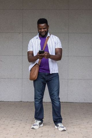 Smiling african man standing using smartphone with crossbody leather bag urban casual style