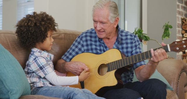 Grandfather and grandchild are sitting on a couch, with the grandfather teaching the child to play an acoustic guitar. They appear relaxed and engaged in the moment. This stock photo can be used for themes related to family bonding, music education, multigenerational relationships, or leisurely domestic activities.