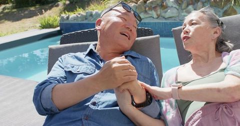 Senior asian couple reclining by pool holding hands enjoying sunny backyard relaxation