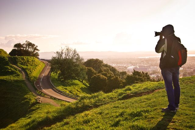 Photographer standing on hillside grass capturing a breathtaking sunset over a winding road. Useful for travel blogs, adventure promotions, landscape photography portfolios, and outdoor exploration themes. Ideal for conveying a sense of discovery, freedom, and the beauty of nature.