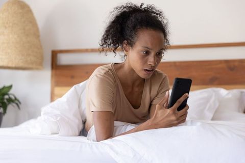 African american woman lying on bed looking shocked at smartphone morning reaction