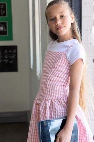 Preteen student wearing pink gingham dress holding tablet near sunlit classroom window