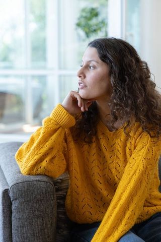 Contemplative woman wearing mustard knit sweater sitting on gray sofa by sunlit window
