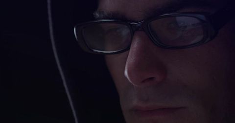 Hooded Man with Reflection on Glasses Focused on Screen in Dark Room