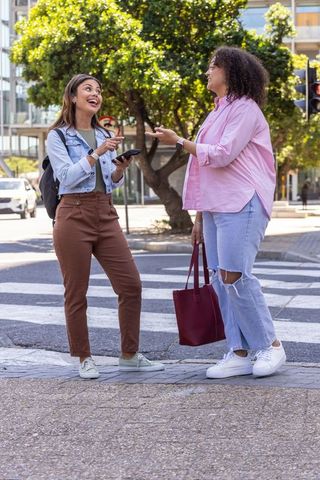 African american friends chatting at city crosswalk with smartphone and tote bag