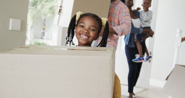 Happy family moving into new home, carrying cardboard boxes while smiling. The young girl in the foreground expresses excitement, making the moment joyful and memorable. Perfect for illustrating topics of homeownership, moving, new beginnings, family togetherness, and happiness in diverse families.
