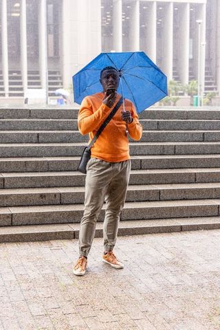 African american man holding blue umbrella checking phone on urban steps in rain commuter portrait