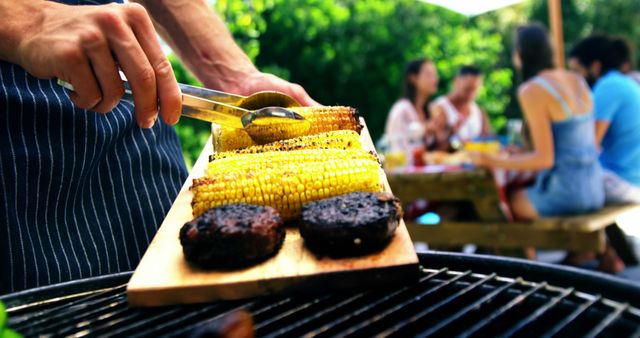 Person grilling corn and burgers on a barbecue in a backyard during a summer gathering. Friends are seen socializing at a picnic table in the background. Perfect for use in articles, blogs, and advertisements related to summer activities, barbecues, cooking, and outdoor social gatherings.