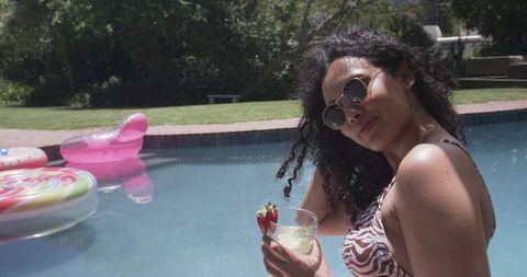 African american woman sipping cocktail by pool wearing sunglasses in sunny backyard