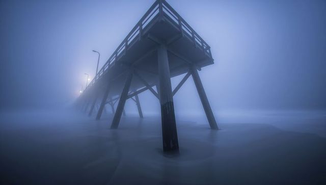 Wooden pier vanishing into dense coastal fog at twilight with glowing lampposts and smooth sea