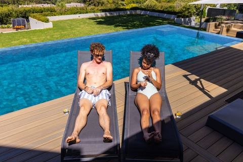 Couple sunbathing on lounge chairs by blue-tiled infinity pool, backyard deck relaxation